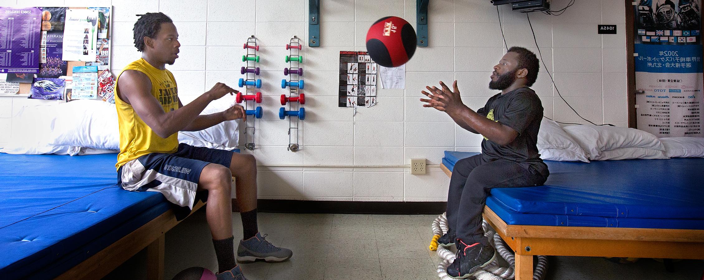 Two people toss a medicine ball while sitting down in a training room.