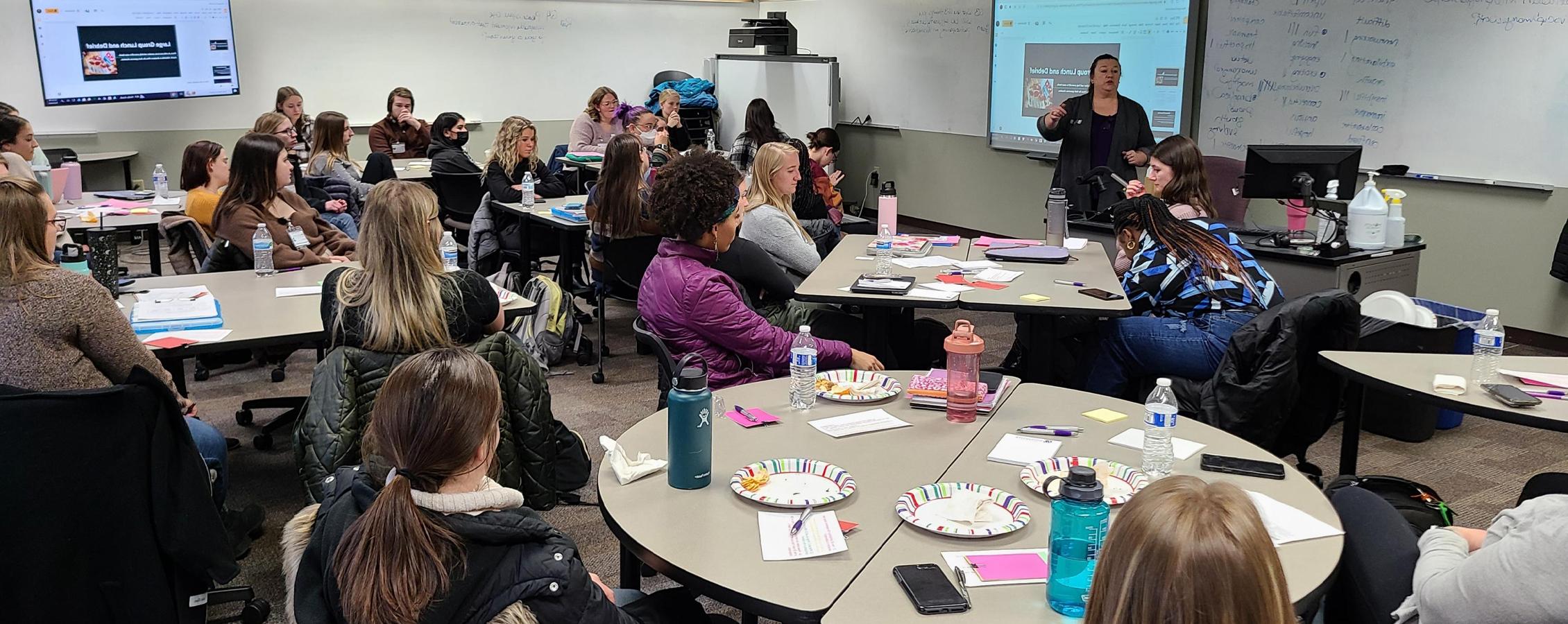 A faculty member speaks to a large group of students in a classroom setting.